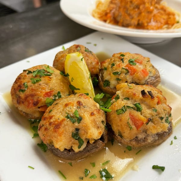 Five stuffed mushrooms with a lemon wedge and parsley, served in a light sauce on a white plate.