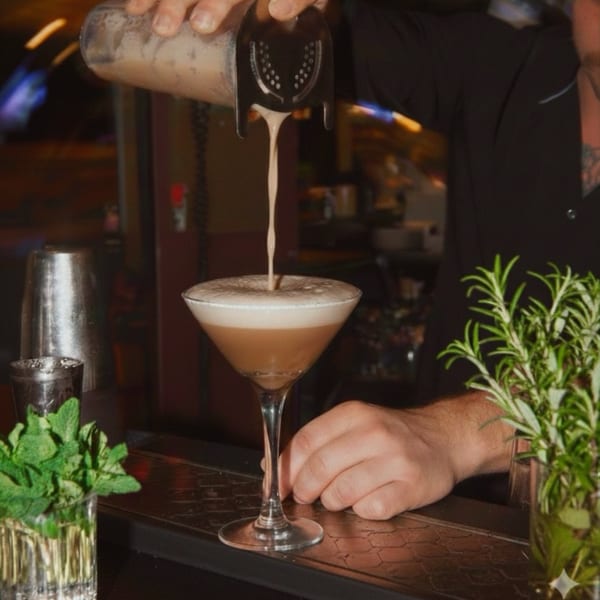 Bartender pouring a creamy brown cocktail from a shaker into a martini glass on a dark bar.