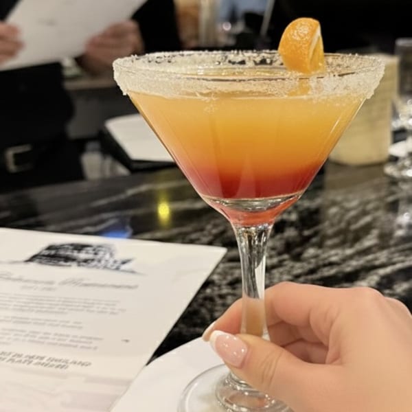 A hand holds a vibrant orange and red cocktail in a sugar-rimmed martini glass with an orange garnish.