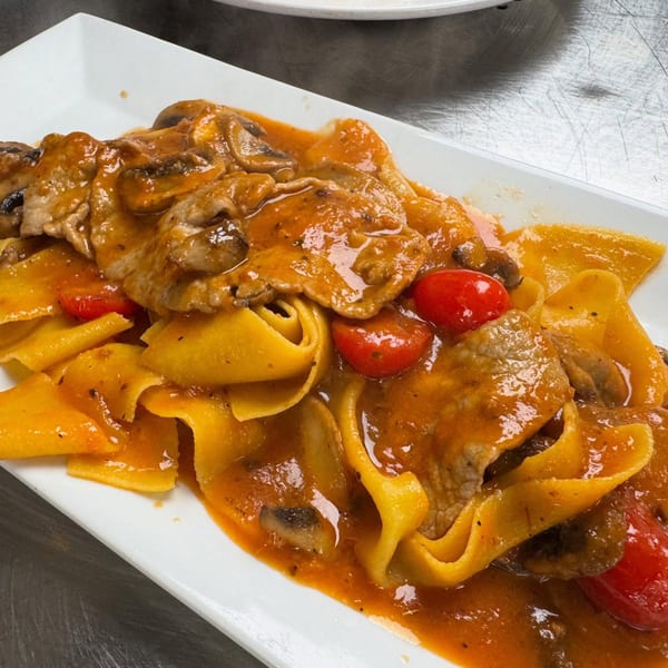 Pappardelle pasta with meat, mushrooms, and cherry tomatoes in a rich red sauce on a white plate.