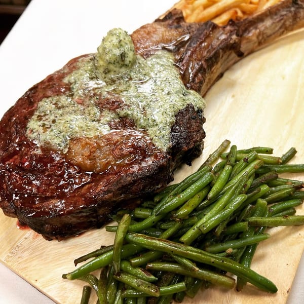 Tomahawk steak with herb butter, green beans, and french fries on a wooden board.
